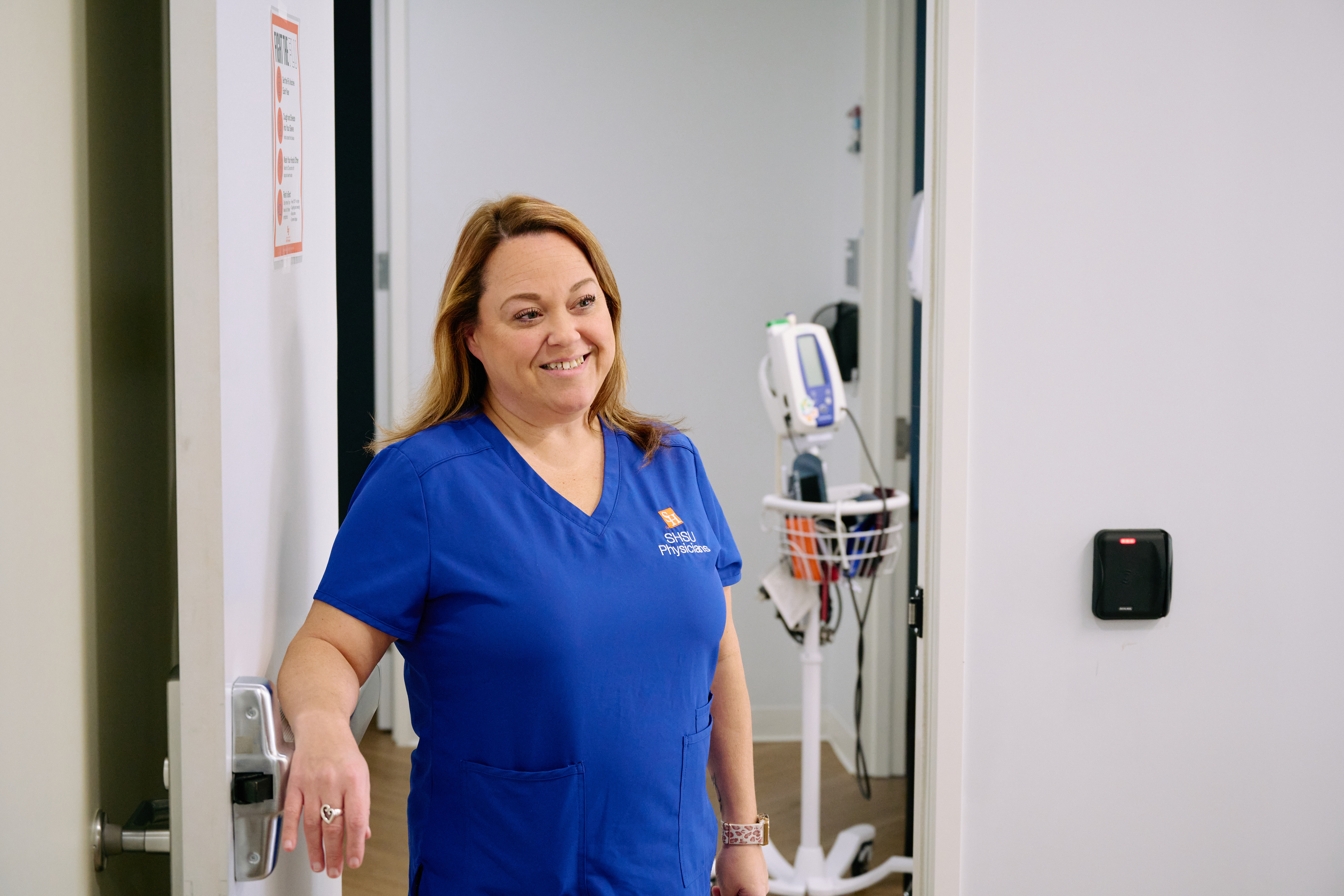 Nurse in blue SHSU Physicians scrubs smiling and waiting for a patient to come into an exam room